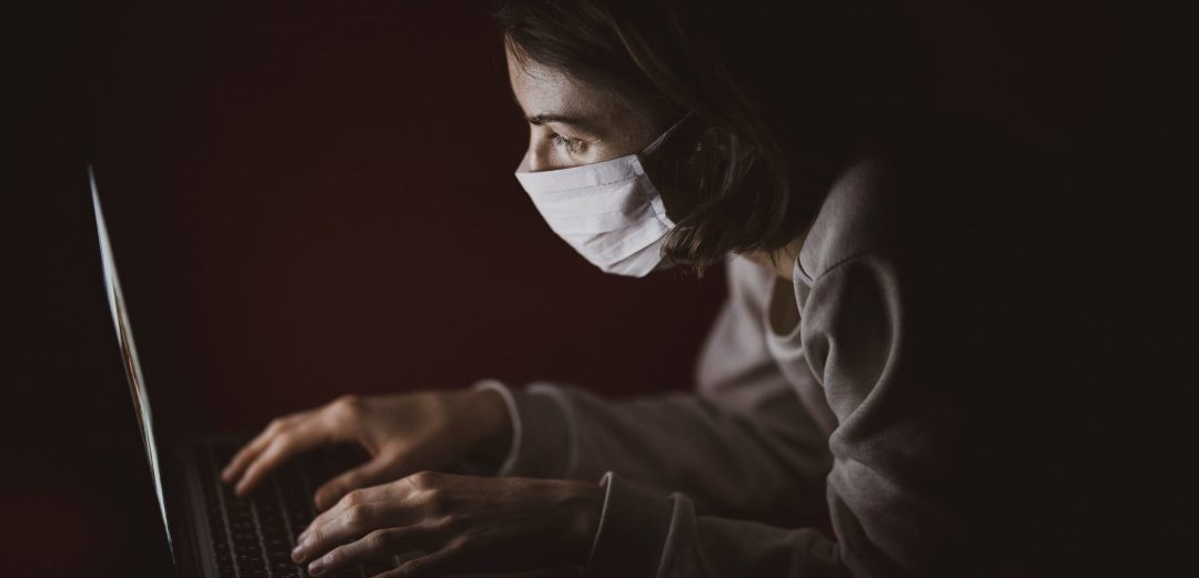 Image of a woman wearing a medical facemask huddled over a laptop. The light from which illuminates her face.